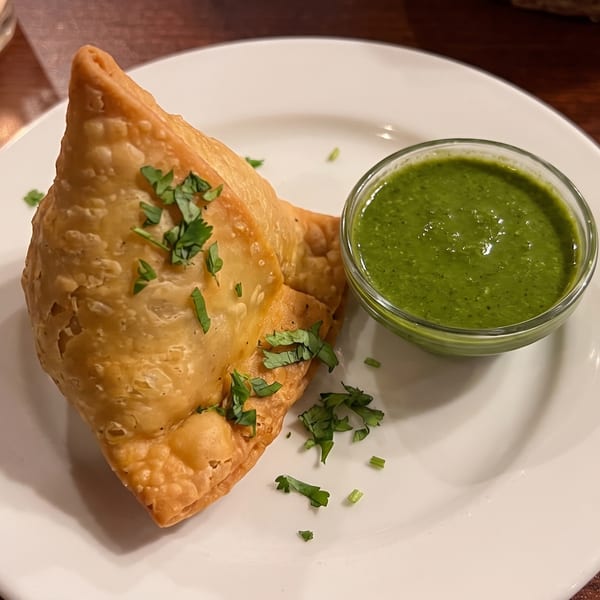 Golden-brown samosa garnished with cilantro, served with a side of green chutney on a white plate.