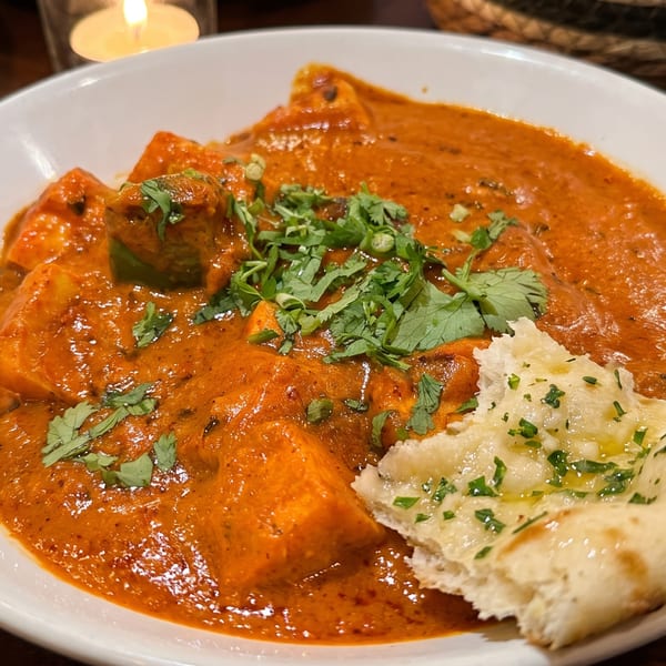 Paneer butter masala with cilantro and a piece of naan bread in a white bowl.