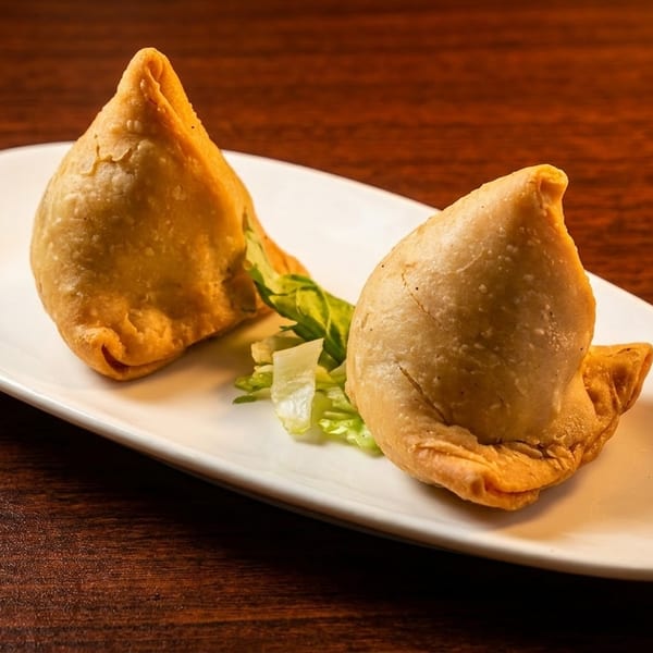 Two golden-brown samosas with a garnish of lettuce on a white plate, on a wooden table.