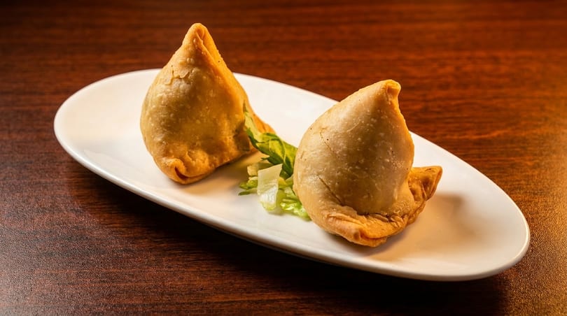 Two golden-brown samosas with a garnish of lettuce on a white plate, on a wooden table.